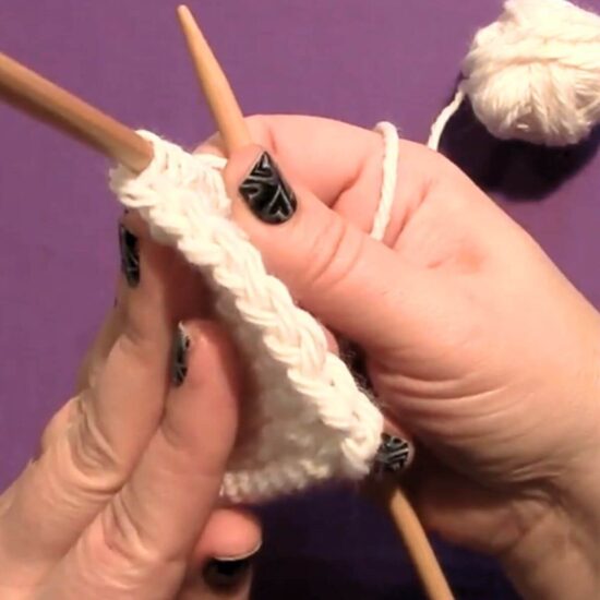 Two white hands showing the chain edge of a piece of knitting with the wood needles still in the knitting. The yarn is very pale pink and the background is purple.