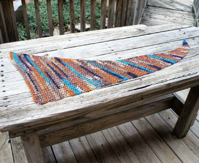 A wood bench on a wood deck. Laying across the bench is a stretched asymmetrical triangular v-stitch crochet wrap with a long, skinny end, wrap is crocheted with multicolor yarn in shades of blues and tans.