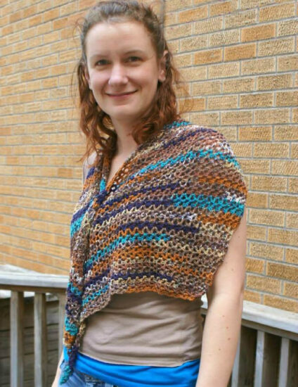 Young woman in front of a brick wall wearing a beige sleeveless top. She's also wearing a stretched asymmetrical triangular v-stitch crochet wrap with a long, skinny end. The wrap is crocheted with multicolor yarn in shades of blues and tans. The side opposite the long point is draped over one shoulder and the long point is draped over the other.