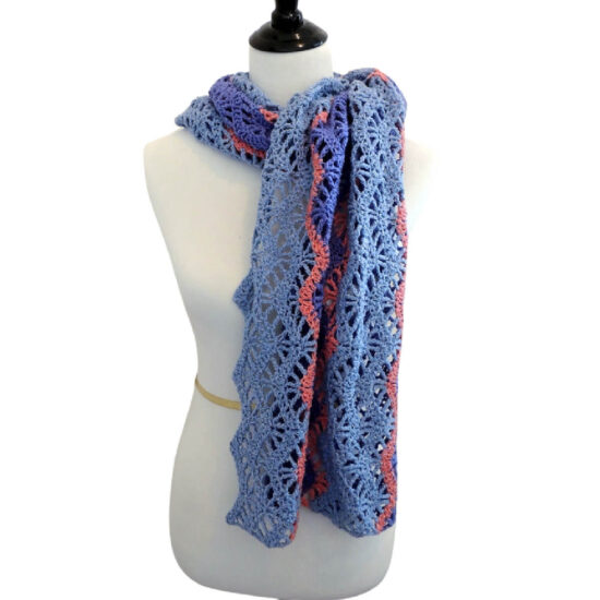 Off-white dress form in a lightweight chevron crochet wrap with alternating rows of solid stitches and lacy open stitches which accentuate the chevrons. The wrap is made with thick strips of light and medium blue yarn separated by thin stripes of coral yarn. The wrap is wrapped around the neck and tied like a scarf.