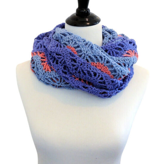 Off-white dress form in a lightweight chevron crochet wrap with alternating rows of solid stitches and lacy open stitches which accentuate the chevrons. The wrap is made with thick strips of light and medium blue yarn separated by thin stripes of coral yarn. The wrap is bunched up and wrapped around the neck like a cowl.