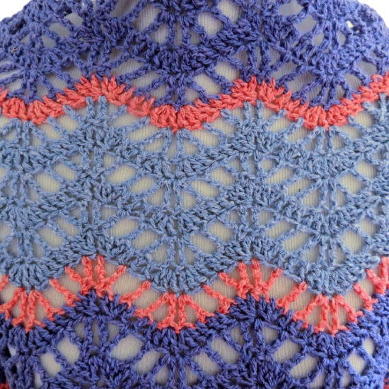 Super close up of the back of an off-white dress form in a lightweight chevron crochet wrap with alternating rows of solid stitches and lacy open stitches which accentuate the chevrons. The wrap is made with thick strips of light and medium blue yarn separated by thin stripes of coral yarn.