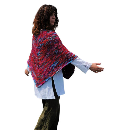 Side view of a young woman with curly hair wearing a white blouse, black pants and a classic poncho with points in the front and back. The poncho is knit with yarn that's mostly scarlet with short lines of blues and pinks. The young woman is turning slightly with her arms out at a 45 degree angle.