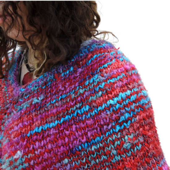 Close up of the front side of a young woman with curly hair wearing a poncho knit with yarn that's mostly scarlet with short lines of blues and pinks.