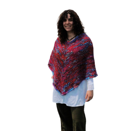 Slightly turned front view of a young woman with curly hair wearing a white blouse, black pants and a classic poncho with points in the front and back. The poncho is knit with yarn that's mostly scarlet with short lines of blues and pinks.