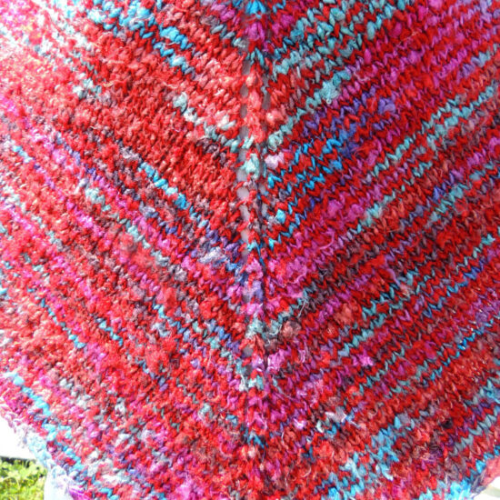 Super close up of the front of a poncho knit with yarn that's mostly scarlet with short lines of blues and pinks.
