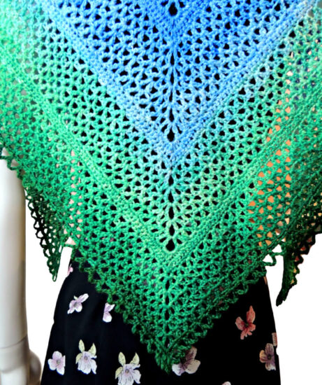 Close up of the back of a mannequin in a black tank top and skirt with a triangular crochet lace shawl with a scalloped chain edge. The shawl changes color from the top center out from dark blue to light blue to light green to dark green.