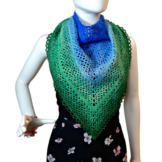 Waist up of a mannequin in a black tank top and skirt with a triangular crochet lace shawl with a scalloped chain edge draped in front with the sides gathered and pulled to the back. The shawl changes color from the top center out from dark blue to light blue to light green to dark green.