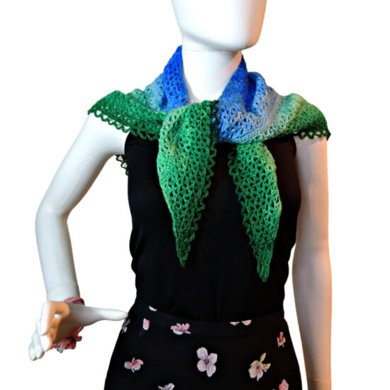 Waist up of a mannequin in a black tank top and skirt with a triangular crochet lace shawl with a scalloped chain edge tied around its neck. The shawl changes color from the top center out from dark blue to light blue to light green to dark green.