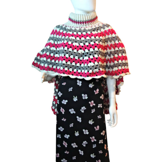 Front view of a mannequin in a black skirt and a poncho made of a large crochet granny stitch circle, the turtleneck of the poncho is about half way from the center to the edge, so that one side of the poncho is much shorter than the other. The poncho is made in stripes of gray, white, and dark pink. The short side of the poncho is in the front.