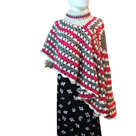 Front view of a mannequin in a black skirt and a poncho made of a large crochet granny stitch circle, the turtleneck of the poncho is about half way from the center to the edge, so that one side of the poncho is much shorter than the other. The poncho is made in stripes of gray, white, and dark pink. The poncho is being worn sideways so the short side drapes off one shoulder and the long side drapes off the other