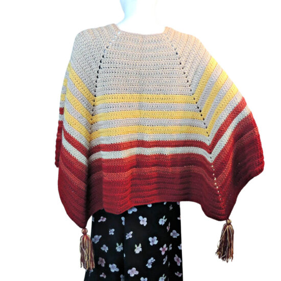 Back view of a white mannequin in a black skirt and a square poncho with one of the straight sides along the back. The poncho is crocheted in yarn starting in tan, then changing in stripes to yellow, rust, then burgundy. Tassels hang off the points.