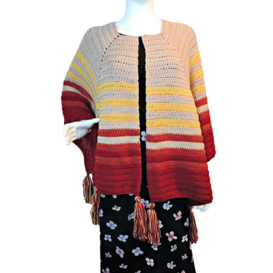 Front view of a white mannequin in a black skirt and an open front poncho that's essentially a square with a slit in the from the center of one side to the center of the square. The slit is in the center front. The poncho is crocheted in yarn starting in tan, then changing in stripes to yellow, rust, then burgundy. Tassels hang off the points.