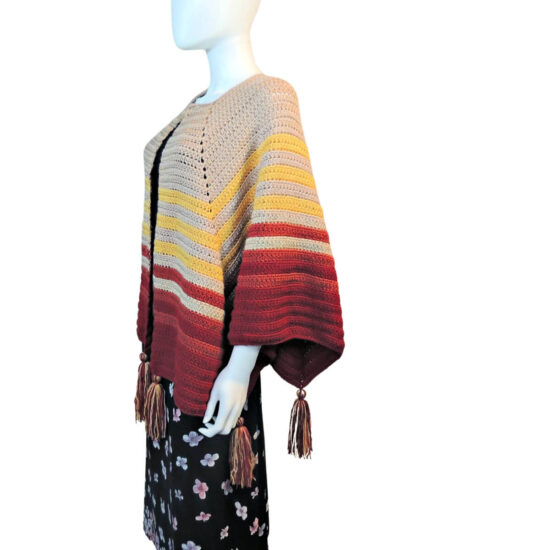 Side view of a white mannequin in a black skirt and an open front poncho that's essentially a square with a slit in the from the center of one side to the center of the square. The slit is in the center front. The poncho is crocheted in yarn starting in tan, then changing in stripes to yellow, rust, then burgundy. Tassels hang off the points.