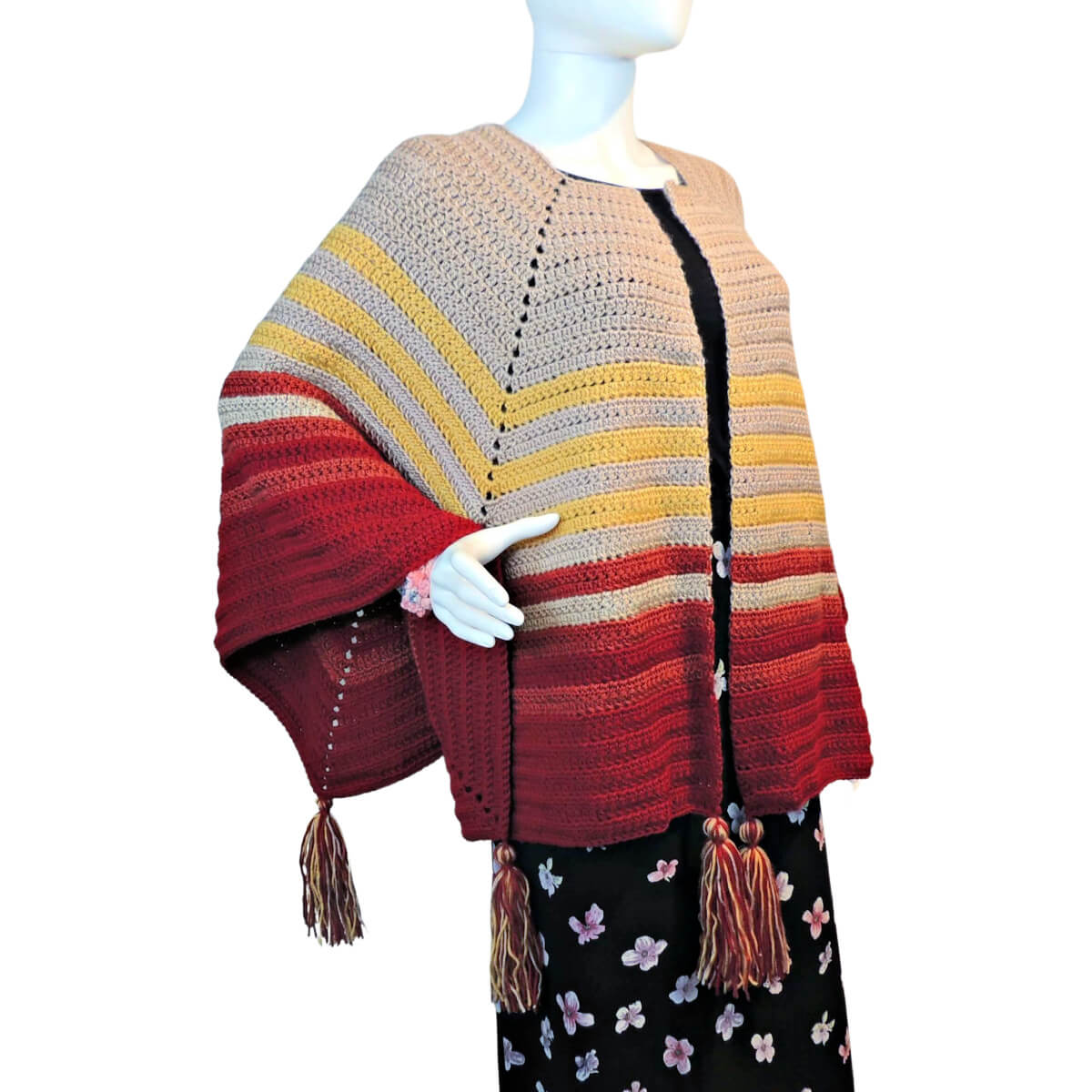 Side front view of a white mannequin in a black skirt and an open front poncho that's essentially a square with a slit in the from the center of one side to the center of the square. The slit is in the center front. The poncho is crocheted in yarn starting in tan, then changing in stripes to yellow, rust, then burgundy. Tassels hang off the points.