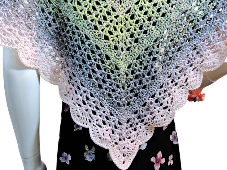 Close up of a triangular crochet shawl with the long center on the the back of a white mannequin. The shawl is lacey and the part we can see changes pastel colors from green to gray to pink.