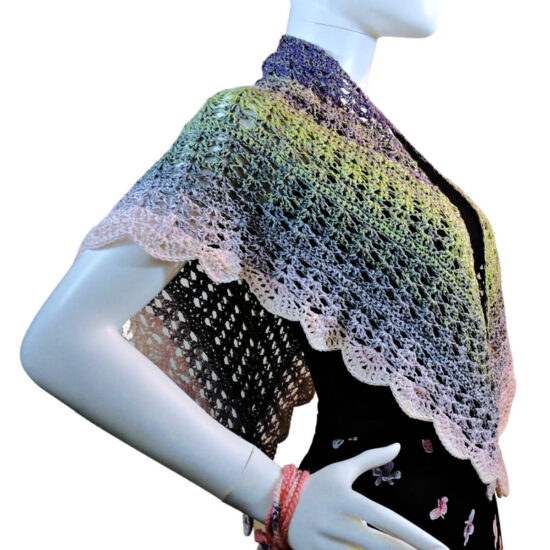 Close up of the side of the torso of a white mannequin wearing a triangular crochet shawl with the long center in back and the two sides draped over the shoulders. The shawl is lacey and changes pastel colors from purple to green to gray to pink.