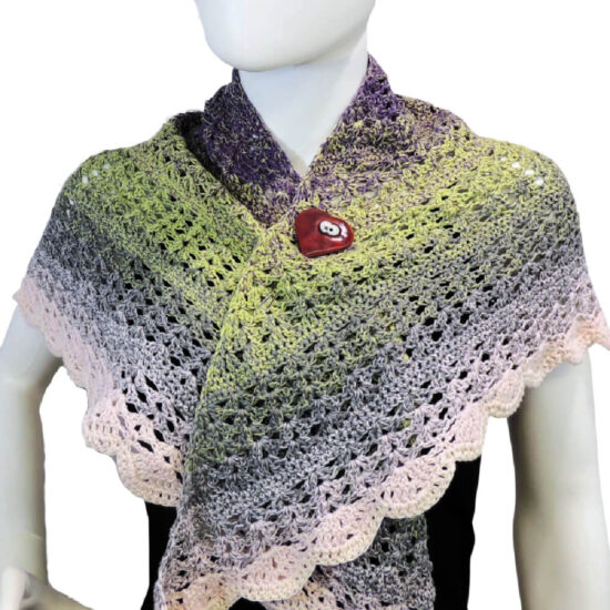 Close up of the front of the torso of a white mannequin wearing a triangular crochet shawl with the two sides draped over the shoulders. A red heart shaped shawl pin with white eyes holds the sided together just under the neck. The shawl is lacey and changes pastel colors from purple to green to gray to pink.