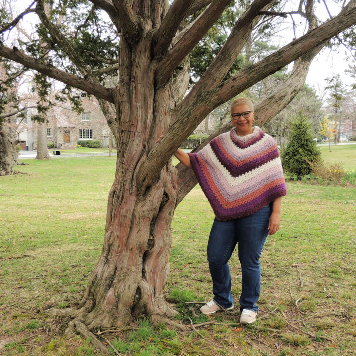 A woman standing next to a tree in a park. She's wearing jeans and a classic 2 point poncho crocheted in fuzzy yarn in stripes from the neck down of cream, lilac, lavender, light rust, purple, and burgundy, repeated so each color is seen 3 times. Her shirt can not be seen beneath the poncho.