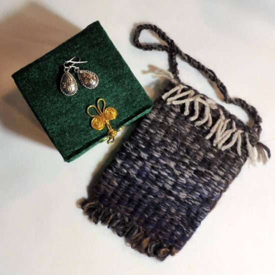 A small woven pouch in navy blue with bits of brown and cream. Next to the pouch is a green velvet box with a gold clasp and silver earrings on top.