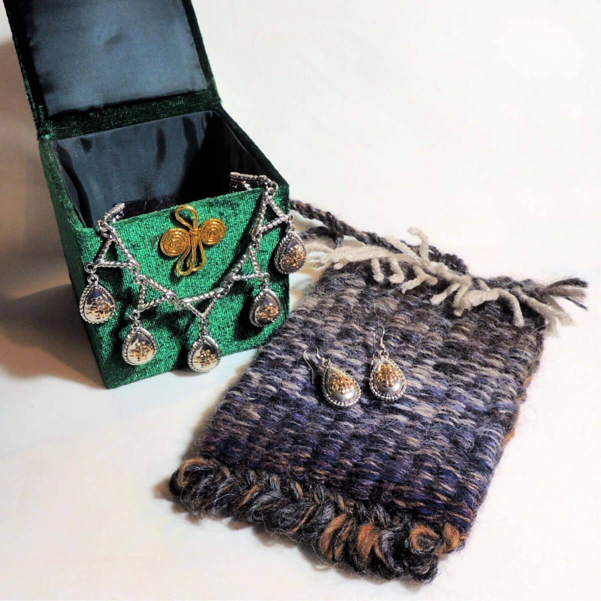 A small woven pouch in navy blue with bits of brown and cream. On top of it is a pair of silver and gold earrings. Next to the pouch is an open green velvet box with black lining, a necklace that matches the earrings hangs out of the box.
