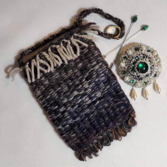 A small woven pouch in dark blue with bits of brown and cream. Next to the pouch are 2 vail pins with green beads at the tops and a large broach with green gems and pearls.