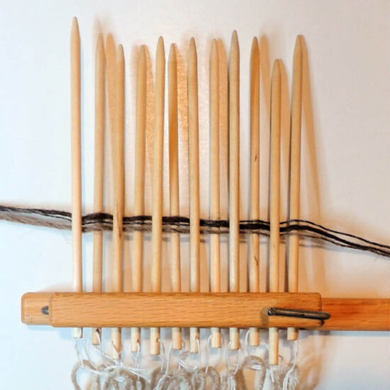 12 pointed wood dowels clamped together in a row just above the bottom. Yarn is threaded through holes in the bottoms of the dowels and more yarn woven through them once above the clamp.