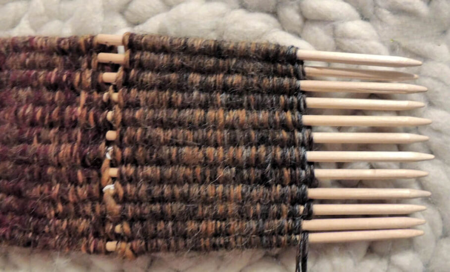 12 pointed dowels laying in a row with yarn woven through them. The weaving continues past the ends of the dowel and off the edge of the image.