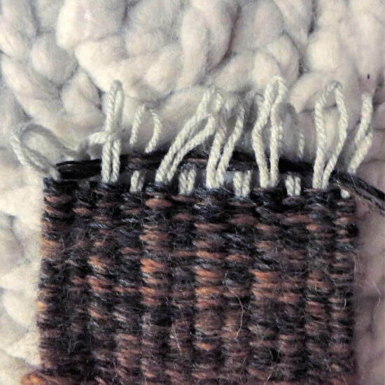 End of dark woven piece with the tops of folded over warp yarn hanging out of the top. The warp loops are in the process of being pulled through each other.