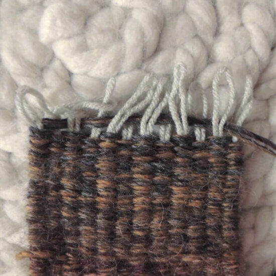 End of dark woven piece with the tops of folded over warp yarn hanging out of the top. The warp loops are in the process of being pulled through each other.