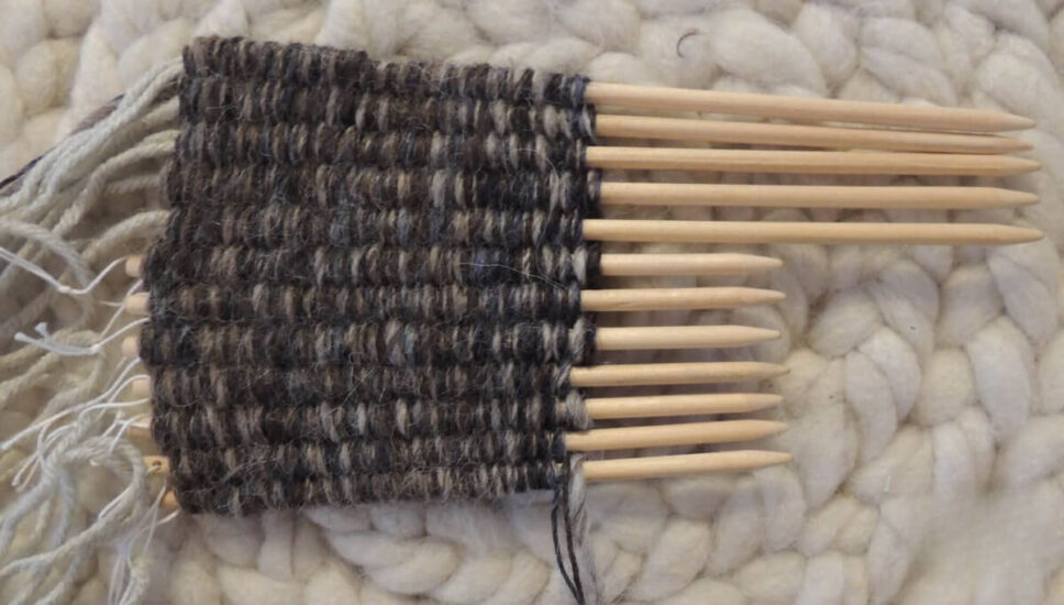 12 pointed dowels laying in a row with yarn woven through them for a few inches. 4 of the dowels have been pulled up longer then the rest. Gray yarn hangs from the bottoms of the dowels.