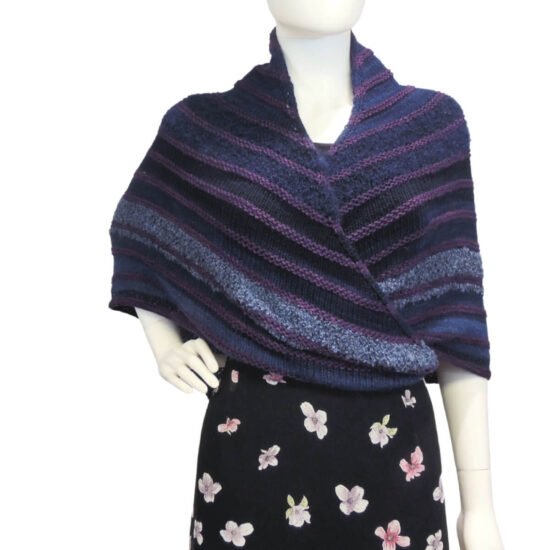 Front view of a white mannequin wearing a black skirt, black tank top, and a large triangular shawl in shades of blue with small raised stripes of purple every few inches, oversized tassels hang off the 3 points. The triangle shawl is a right angle with 2 equal sides, the center of the long side is at the back of the neck and wraps one end at a time to the front then around the back at the waist.