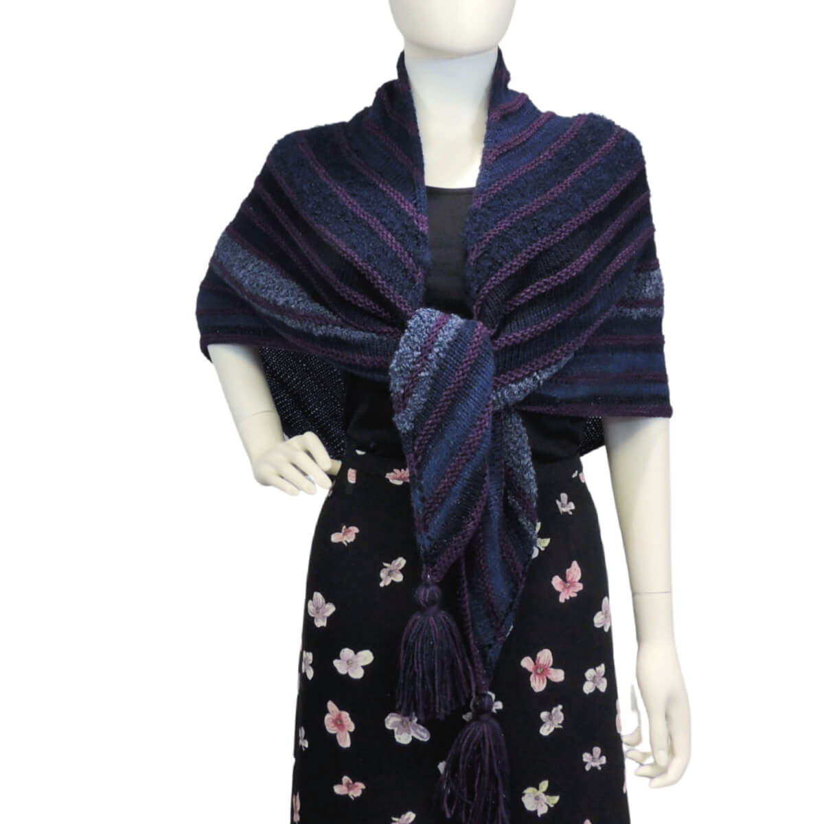 Front view of a white mannequin wearing a black skirt, black tank top, and a large triangular shawl in shades of blue with small raised stripes of purple every few inches, oversized tassels hang off the 3 points. The triangle shawl is a right angle with 2 equal sides, the center of the long side is at the back of the neck and wraps to the front where it's tied together just above the waist.