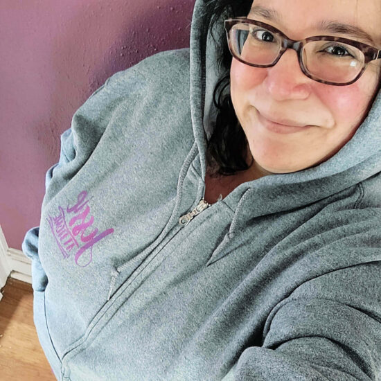 Image of a woman (Jessie) in a gray zip hoodie with a Jessie At Home logo on it.
