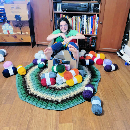 Image of a child (Vada, 13) sitting in a box full of yarn with yarn all over the rug and floor around the box.