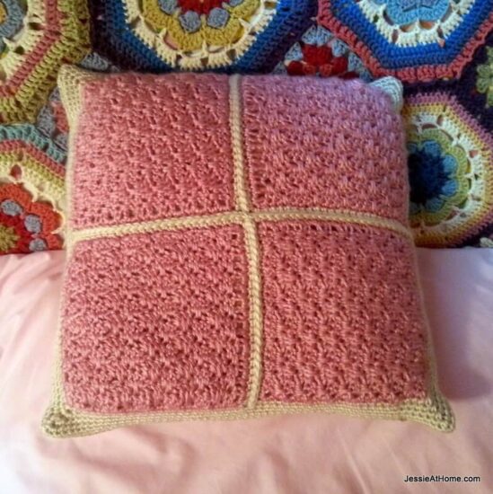 Image of a square pillow with a pink crochet top with gold crochet trim on a pink sheet and multicolored crochet afghan.