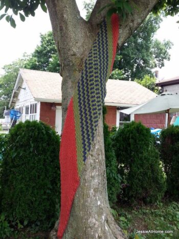 A green, blue, and salmon crochet wrap hanging from a tree with bushes and houses in the background