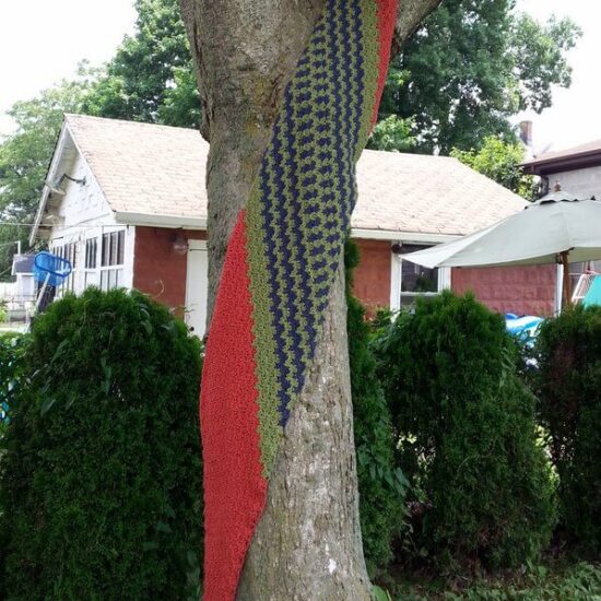 A green, blue, and salmon crochet wrap hanging from a tree with bushes and houses in the background