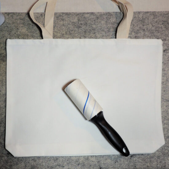 A photo of a plain white tote bag laying flat on a gray background, with a link roller on top of it.