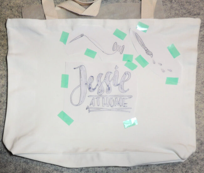 A photo of a plain white tote bag laying flat on a gray background, with various colored art graphics taped face down on it.