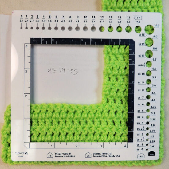 A white square plastic gauge ruler with a 4 inch square opening inside. The ruler is on top of a green piece of crochet that goes across the bottom and up the right side of the photo. Under the crochet is a piece of paper with hand written "4 inches = 14 stitches"