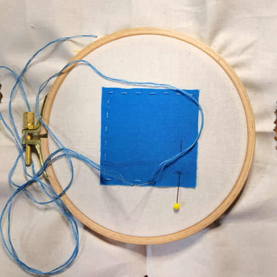 Square of off white fabric stretched in an embroidery hoop with a blue fabric square partially stitched down with a blue running stitch. The needle and embroidery floss are still attached.