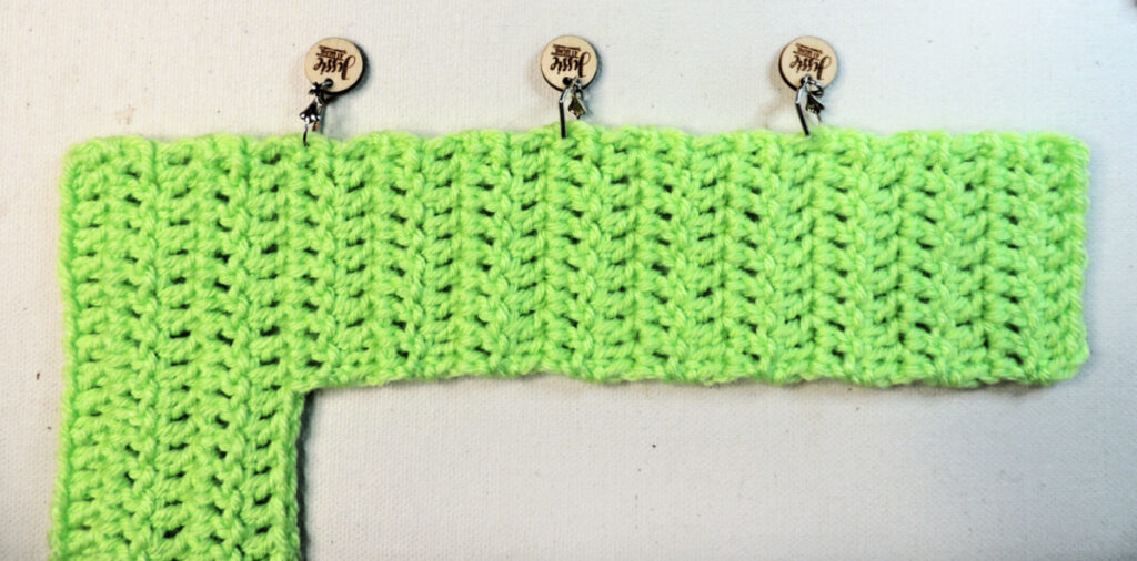 An "L" shape made of green crochet on its side. On the long side are 3 stitch marker dividing it in fourths.
