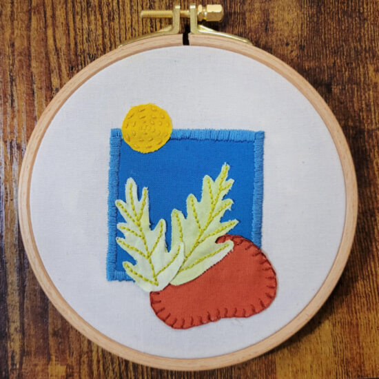 An embroidery hoop with off white fabric stretched in it. On the fabric is a blue square stitched down with blue satin stitch, a yellow circle stitch almost on the top left corner of the square with a running stitch in concentric circles, a rust bean-shape stitched with a mattress stitch on the bottom right corner of the square. Inside the square to the left of the bean are 2 pale green leaves stitched down with backstitch veins.