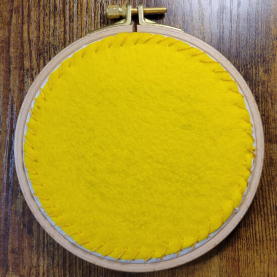 Wood embroidery hoop with a yellow felt circle stitched to the off white fabric stretched in the hoop, so that nearly none of the fabric shows.