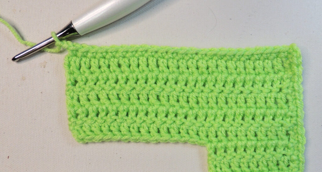 An "L" shape made of green crochet upside down. The bottom of the "L" is on top and at the top left corner is a crochet hook attached to the unfinished border.