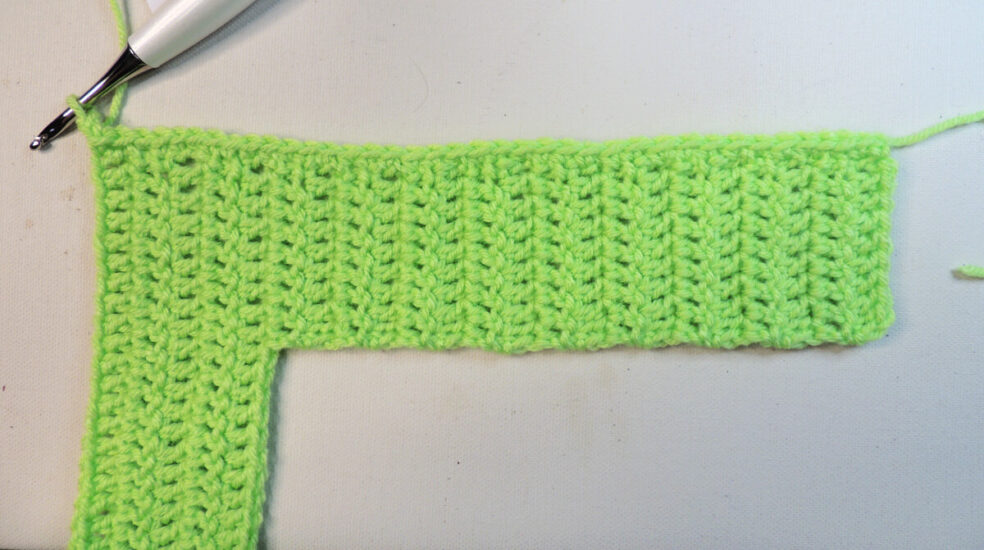 An "L" shape made of green crochet on its side. The long edge is on top and at the top left corner is a crochet hook attached to the unfinished border.