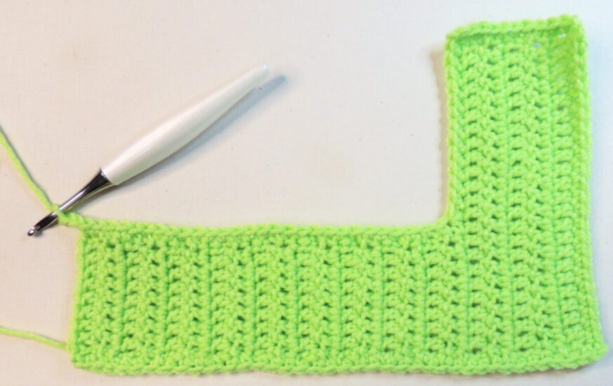 An "L" shape made of green crochet with the long edge at the bottom. At the top left corner of the long edge is a crochet hook attached to the unfinished border.