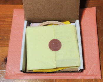 A coral shipping padded envelope on top of a wood table. On top of the envelope is an open thing white box with yellow tissue pattern wrapped around the contents and sealed with a brick colored sticker.