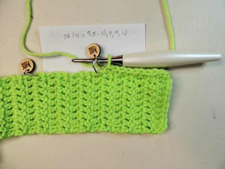 A green rectangular piece of crochet that continues off the left of the image. On the top of the part we can see are 2 stitch markers and a crochet hook about a third of the way across making a border. The crochet sits on a small square piece of paper with some math on it.